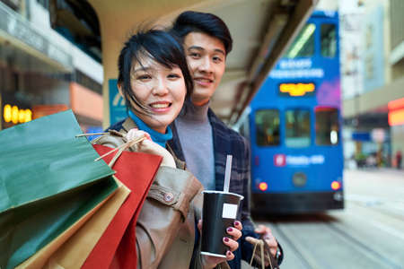 happy young asian couple waiting for bus in modern city with shopping bags in handの写真素材