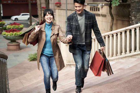 young asian couple carrying shopping bags holding hands walking on steps, happy and smilingの写真素材