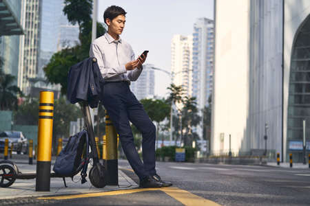 young asian businessman standing next to his electric scooter on street in downtow of modern cityの写真素材