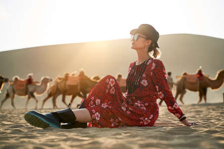 asian woman in red dress sitting in desert looking at view with caravan of camels and huge sand dune in backgroundの写真素材