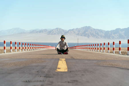 asian woman tourist sitting in the middle of an empty open road with lake and rolling mountains in background, leg crossed.の写真素材
