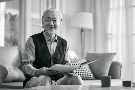 happy asian old man sitting on couch at home reading a book looking at camera smiling, black and whiteの写真素材