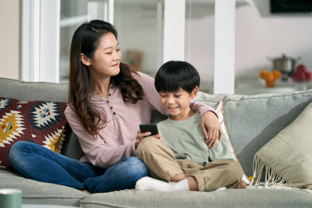 happy asian mother and son sitting on family couch looking at cellphone photos togetherの写真素材