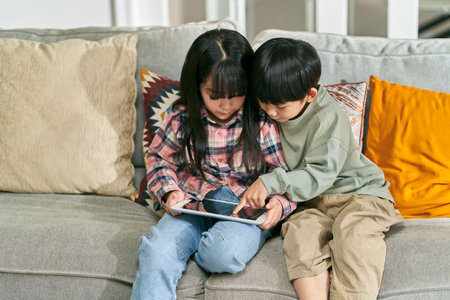 two asian children brother and sister sitting on family couch at home playing computer game using digital tabletの写真素材