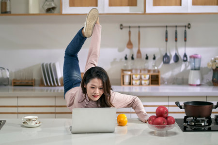 young asian business woman female freelancer stretching legs while working in kitchen at home using laptop computerの写真素材