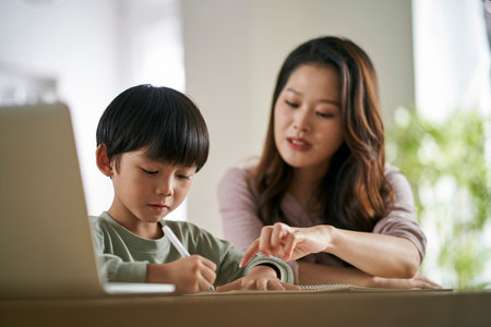 young asian mother sitting at table at home helping son with his study
