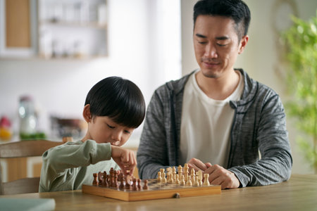 asian father and son sitting at kitchen table at home playing chessの写真素材