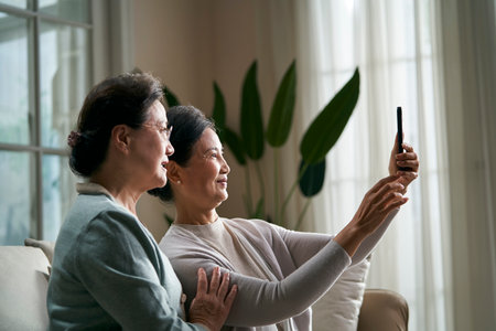 two happy senior asian women sitting on couch at home taking a selfie using cellphone happy and smilingの写真素材