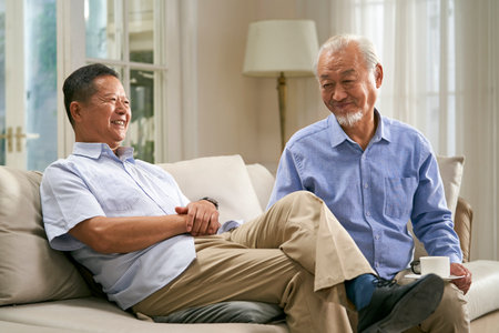 two happy senior asian men sitting and chatting on family couch at home with wives in backgroundの写真素材