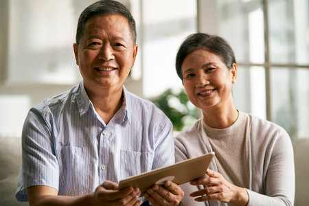 happy senior asian couple sitting on couch at home using digital tablet togetherの写真素材
