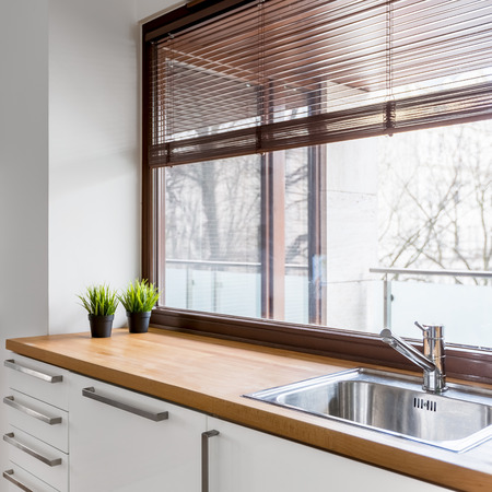 White kitchen with wooden countertop, silver sink and big window with blindsの写真素材
