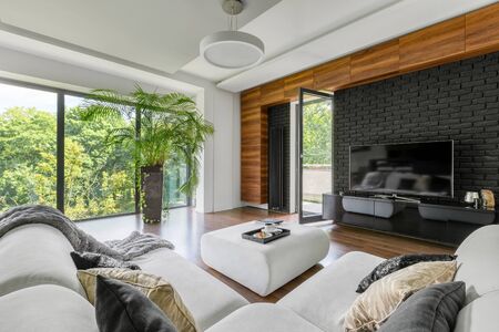 Big white corner sofa with pillows and ottoman for coffee table in elegant living room with big windowsの写真素材