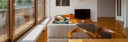 Panorama of sunny living room with big windows, wooden floor, gray corner sofa and tv