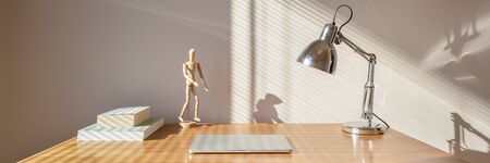 Panorama of simple wooden desk with books, computer, lamp and wooden man and sunlight in roomの写真素材