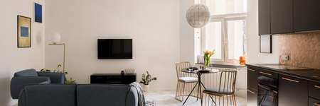 Panorama of small and designed apartment with elegant living room area, stylish dining table with new golden chairs and modern black kitchen in one roomの写真素材