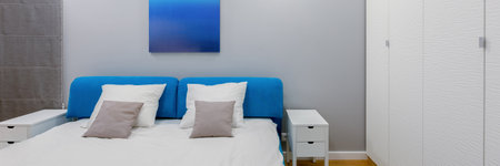 Panorama of modern bedroom with big bed with blue headboard and art, white wardrobe and bedside tablesの写真素材