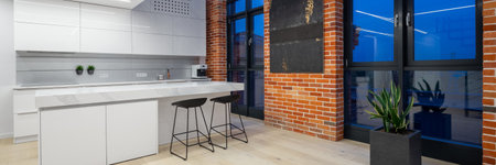 Panorama of stylish kitchen with white furniture and led lights in loft apartment with big windows and brick walls, at nightの写真素材