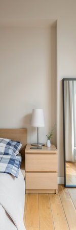 Vertical panorama of simple wooden bedside table with lamp next to bed and long mirror in black frameの写真素材