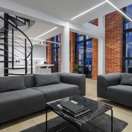 Simple living room with gray sofa and armchair and modern coffee table in open plan loft apartment with brick walls, big windows and spiral stairsの写真素材