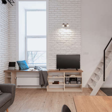 Television screen on simple wooden sideboard in room with narrow window with windowsill seat and white brick on the wallsの写真素材