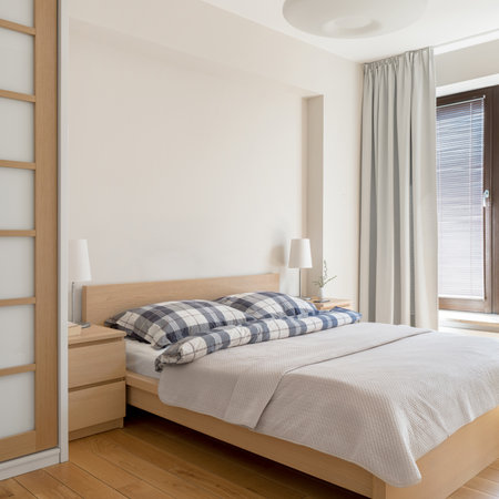 Comfortable, big bed in bright bedroom with wooden furniture and floor and window with blinds behind curtainsの写真素材