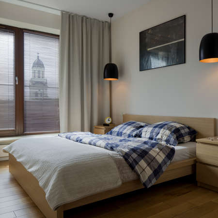 Modern bedroom with big window with decorative blinds and curtains and two black pendant lamps above bedside tablesの写真素材