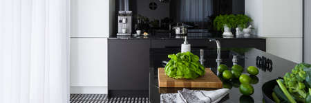 Panorama of green food in luxurious kitchen with black and white furniture, big window behind white curtains and long kitchen island with black countertop and sinkの写真素材