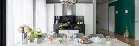Panorama of beautiful decorated and elegant dining table with stylish chairs in apartment with open black and white kitchen and green wallの写真素材