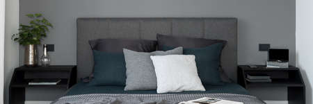 Panorama of simple and elegant bedroom with gray wall and comfortable bed with gray, upholstered headboard and two, black bedside tables with decorationsの写真素材
