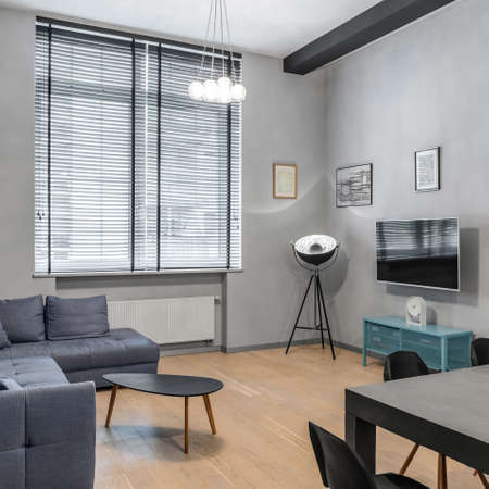 Big window in stylish living room with simple corner sofa, black coffee table, modern lamp on tripod and blue sideboard under television screenの写真素材