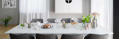Panorama of stylish decorated white dining table with eight, gray and elegant chairs in room with big windows behind white curtainsの写真素材