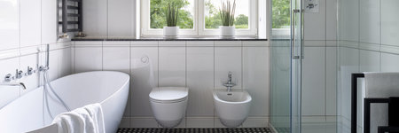 Panorama of spacious and elegant bathroom with white tiles, window, big bathtub, shower cabin and bidet next to toiletの写真素材