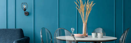 Panorama of fancy dining table with many decorations on marble style countertop with four transparent chairs in elegant room with teal blue wall with moldingの写真素材