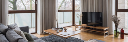Panorama of elegant and bright living room with many windows, comfortable sofa and wooden coffee table with nice decorations and big television screen on wooden sideboardの写真素材