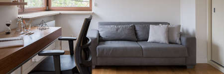 Panorama of comfort zone in simple home office room with window, gray, comfortable couch and wooden desk with black chairの写真素材