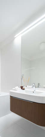 Vertical panorama of mirror wall above modern, white washbasin with wooden drawer in new bathroom with white tiles and led lightingの写真素材