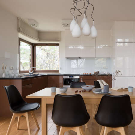 Big, wooden dining table with black chairs in spacious kitchen with white and brown furniture and windowの写真素材