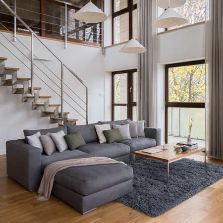 Comfortable, big, corner sofa, wooden coffee table, many windows and stairs in stylish living roomの写真素材
