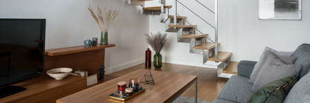 Panorama of modern wooden stairs with silver railing in small and stylish living room with wooden furniture, tv and gray sofaの写真素材