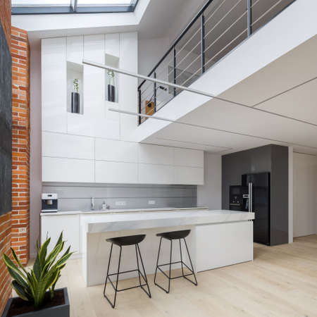 New kitchen with elegant white furniture in stylish loft apartment with mezzanine floor and brick wallsの写真素材