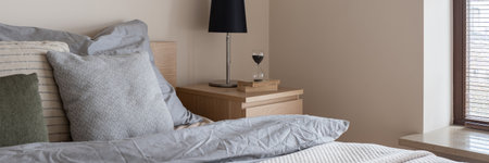 Panorama of black lamp on wooden bedside table with books and hourglass next to comfortable bed in small bedroom with windowの写真素材