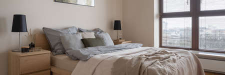 Panorama of comfortable and stylish bedroom with cozy bed, big window with blinds and two wooden bedside tables with black lamps and decorationsの写真素材