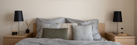 Panorama of comfortable bed with nice pillows, quilt and bedclothes with two simple wooden bedside tables with black lamps and decorations in bright bedroomの写真素材