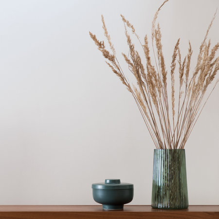 Close-up on green, glass vase with dried grass and green decorative container on wooden shelf with...