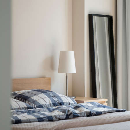 Simple, white lamp on wooden bedside table near bed and mirror in black frameの写真素材