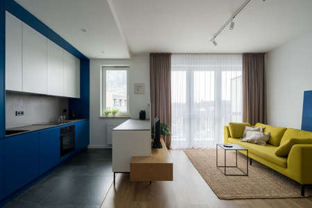 Contemporary apartment interior with modern designed kitchen with blue cupboards open to stylish living room with yellow couchの写真素材
