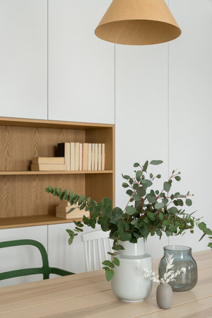 Stylish decorations, vase and plants on wooden dining table in room with white cabinets and wooden shelfの写真素材