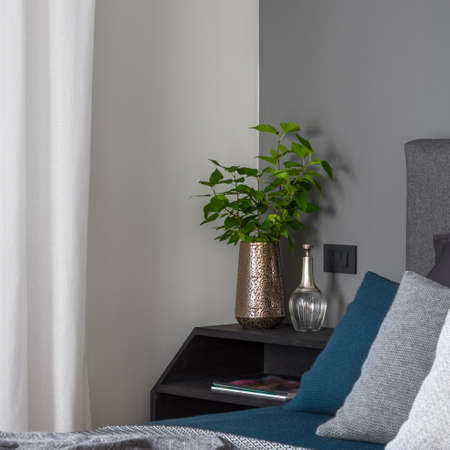 Close-up on stylish golden vase with green plant and decorations on simple, black nightstand next to cozy bed in bright bedroomの写真素材