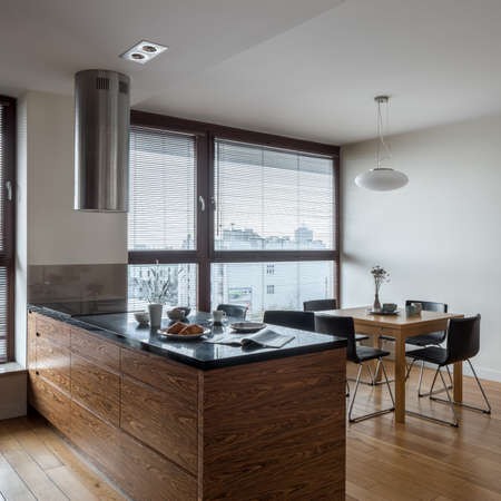 Stylish and bright kitchen with big window, wooden floor, nice kitchen island with black countertop and with wooden dining table with black chairs under pendant lampの写真素材