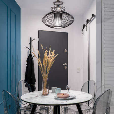 Small studio apartment with stylish dining table with transparent chairs under black, pendant lamp open to white entryway with dark doors and black clothes hangerの写真素材